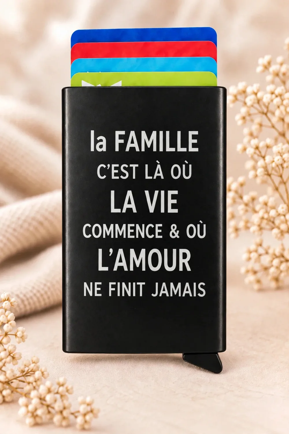 Porte-cartes personnalisé en métal noir, verso gravé d'une citation sur la famille et l'amour, cartes colorées visibles, sur fond de tissu beige et de fleurs
